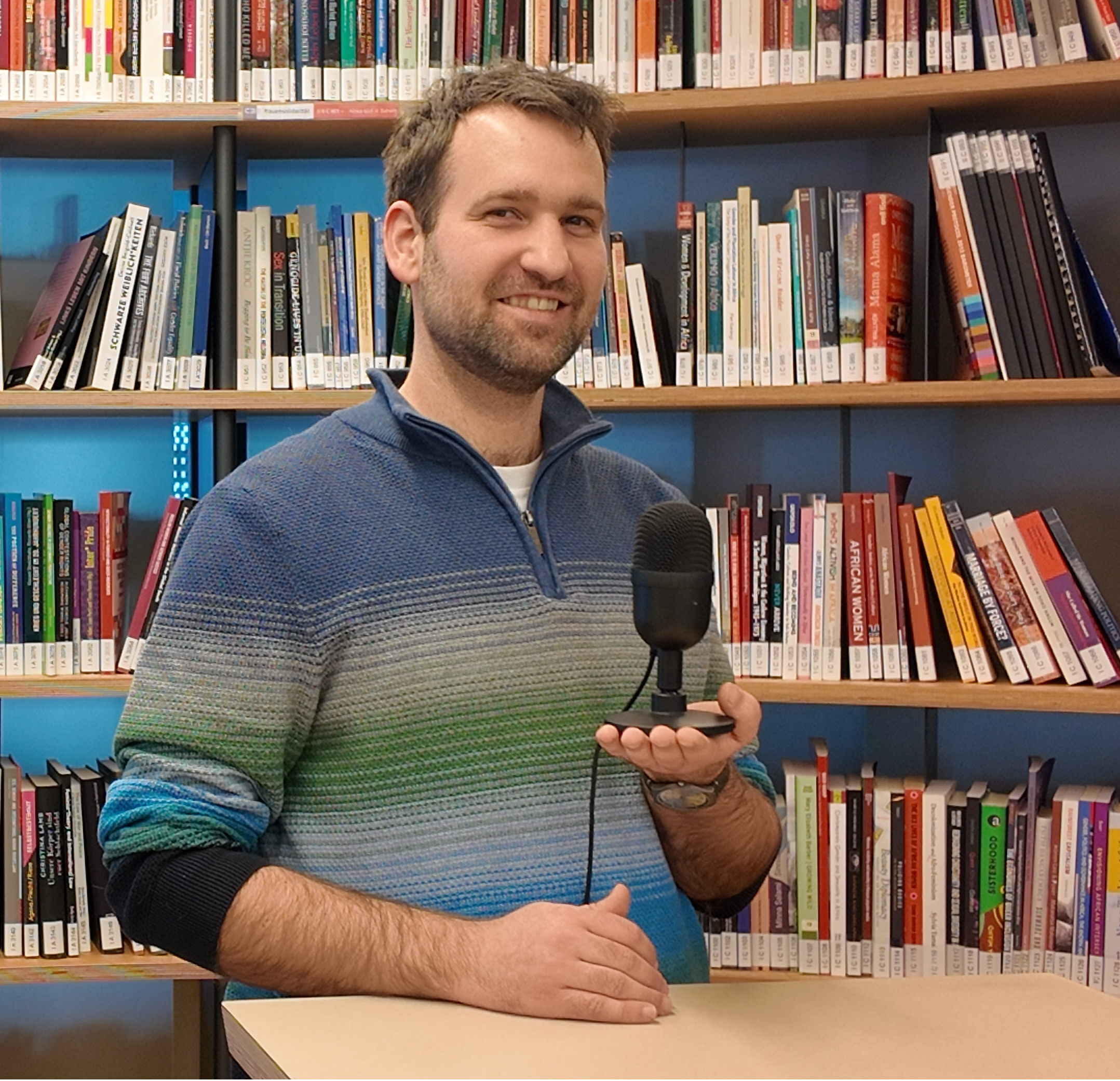 Portrait des neuen Radiohosts mit einem Mikrofon in der Hand. Stehend vor einem Bücherregal.